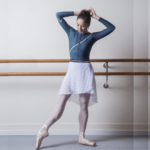 Femal dancer in leotard and short white repertoire skirt pointing foot standing in front of ballet barre