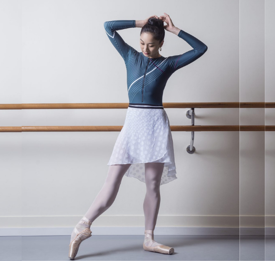 Femal dancer in leotard and short white repertoire skirt pointing foot standing in front of ballet barre