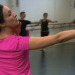 Female adult ballet teacher demonstrating extending her arm to the side to her adult ballet students in teh studio
