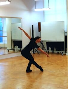 Female adult ballet teacher in a dance studio in a deep lunge reaching down towards her extended front leg.
