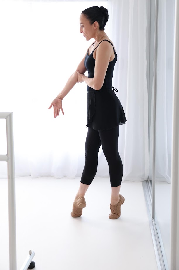 Jane Casson in a white dance studio gracefully poses in a black leotard, skirt, and leggings, with tan dance shoes. She stands on demi pointe, in fourth position, with arms extended in mid demonstration of proper form.