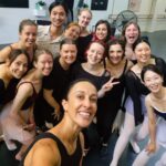 A joyful group of ballet dancers pose for a selfie in a dance studio. They're wearing a mix of black and pastel dancewear, with smiles and cheerful expressions.