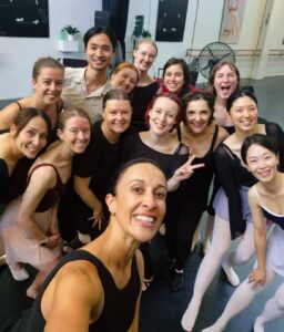 A joyful group of ballet dancers pose for a selfie in a dance studio. They're wearing a mix of black and pastel dancewear, with smiles and cheerful expressions.