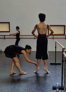 A ballet instructor corrects a male dancer's foot position, one arm extended, in a studio with a barre. A focused atmosphere.